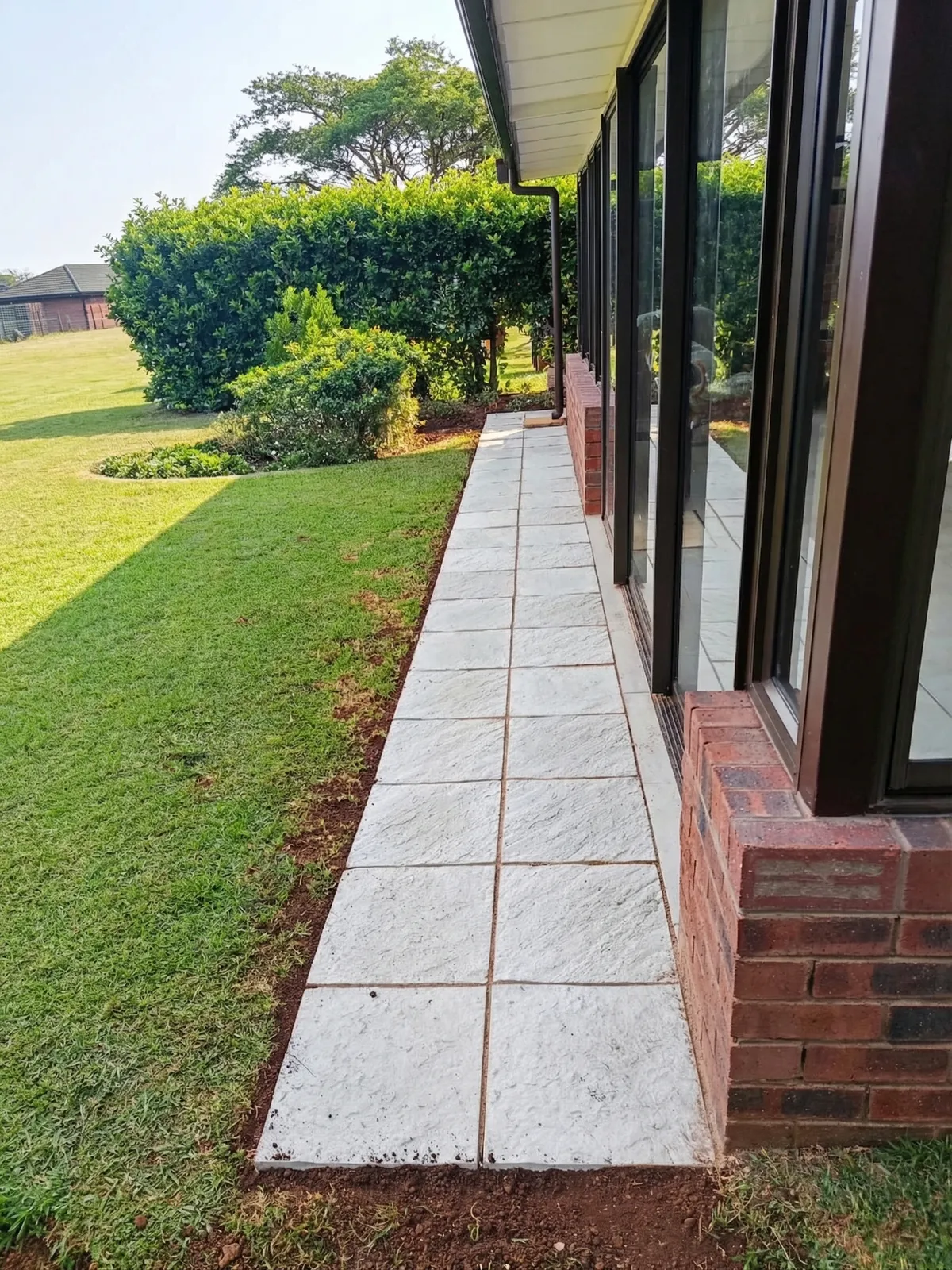 A newly installed walkway made of light grey, textured square pavers running alongside a house with a neatly manicured green lawn.